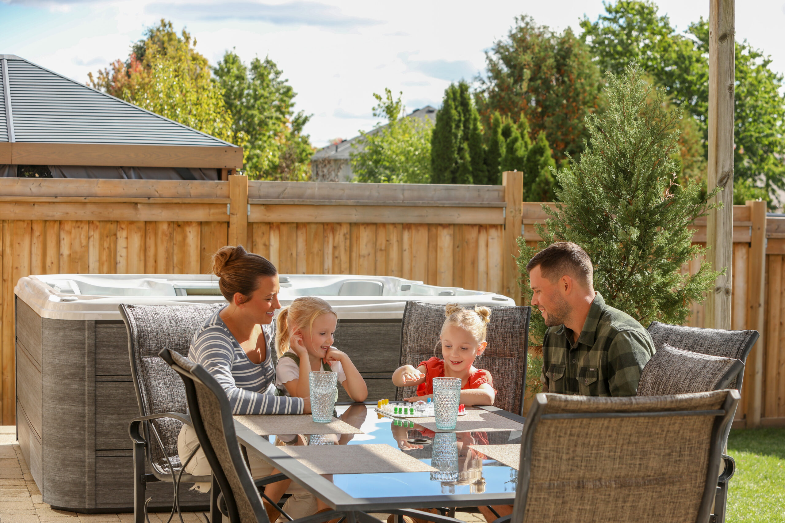 Wide lifestyle photograph of a family enjoying a Sundance 780 Series Chelsee hot tub with a Platinum acrylic shell and Vintage Oak cabinetry, beautifully integrated alongside an outdoor dining table in a luxury garden setting.