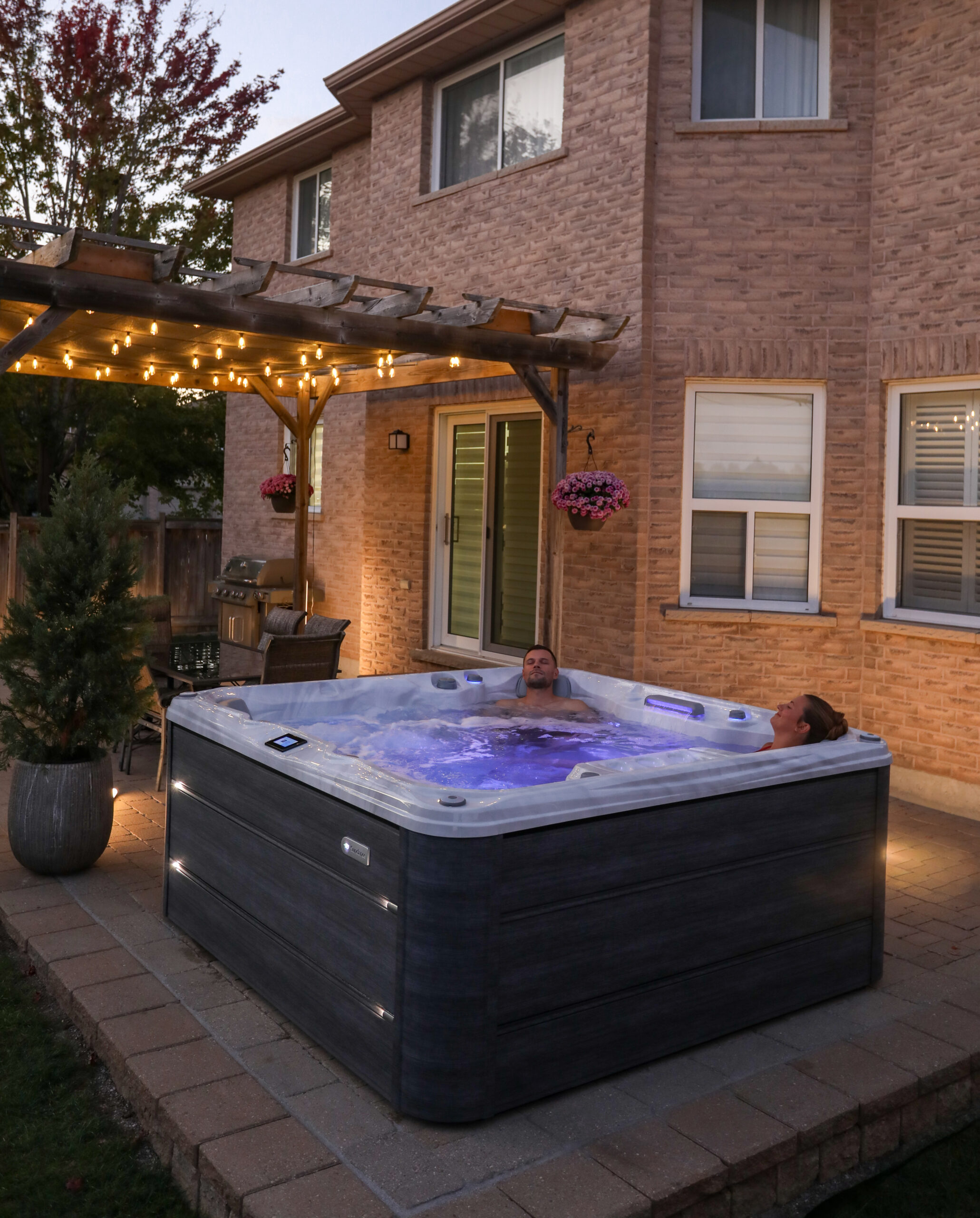 Lifestyle photograph of a couple relaxing together in a Sundance 780 Series Chelsee hot tub featuring a Platinum acrylic shell and Vintage Oak cabinetry, set within a luxury evening garden design.