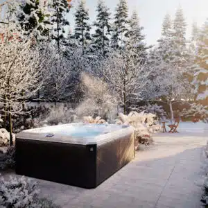 Scenic winter lifestyle photograph of a premium Sundance hot tub steaming in a snow-covered garden setup with frosty trees in the background.