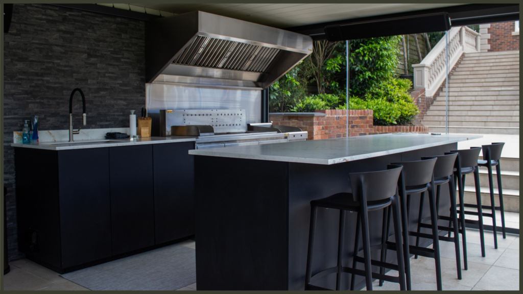 A premium all-weather outdoor kitchen with high-end stainless steel appliances, sheltered beneath a modern black aluminium louvred pergola with its roof closed against rainy British weather.