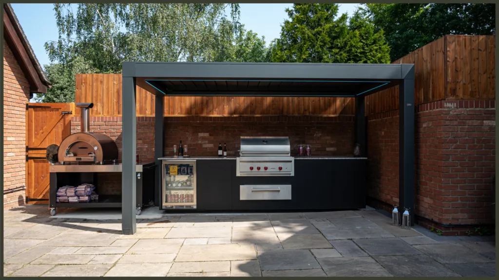 A luxury completed backyard project in Hornchurch, Essex, showcasing a premium built-in outdoor kitchen with granite worktops and an architectural black aluminium louvred pergola over a contemporary dining area.